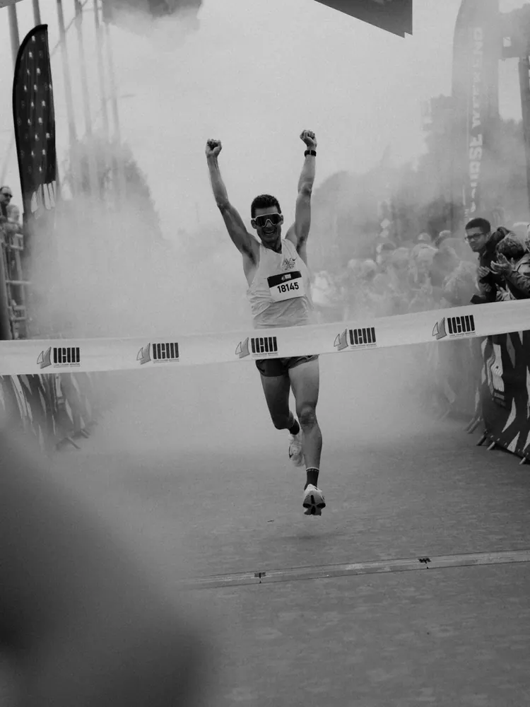 Atleet komt juichend door de rook over de finish, in zwart-wit